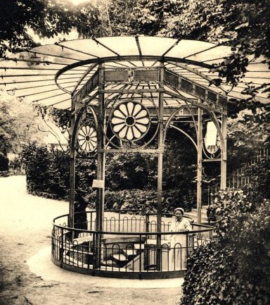 Kiosque en métal avec un toit en verre, entouré de verdure. Atmosphère vintage.
